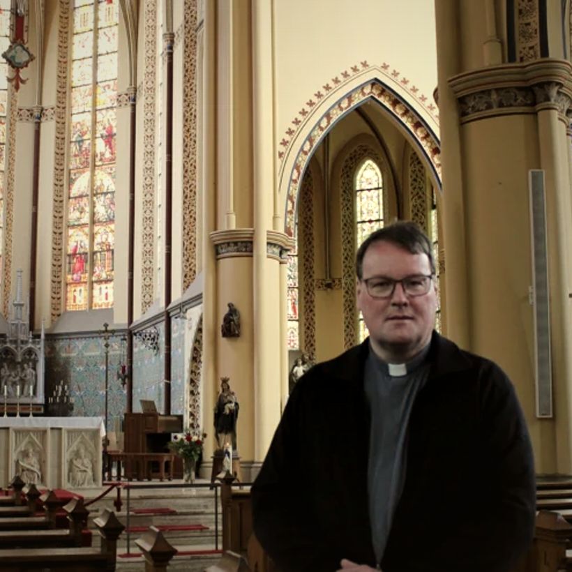 Marc Oortman in priesterboord, staand in de kerk met hoge gewelven en glas-in-loodramen achter hem.