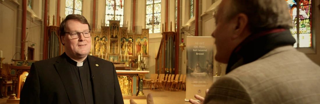 Marc in gesprek met een bezoeker, voor het verguld altaar met brandende kaarsen.