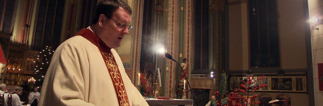 Marc bij de lessenaar tijdens een viering, in het warme licht van kaarsen.